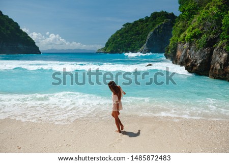 Back view of Woman in bikini walking on beach with sea rocks and turquoise ocean, blue sky. Atuh beach, Nusa Penida island, Bali, Indonesia. Tropical background and travel concept