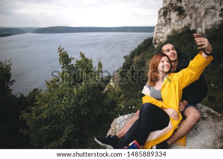 Defocused happy hipster couple taking selfie on phone on top of rock mountain with beautiful view on river. Focus on tree. Tourist couple hugging on windy cliff and making photo. Travel and wanderlust
