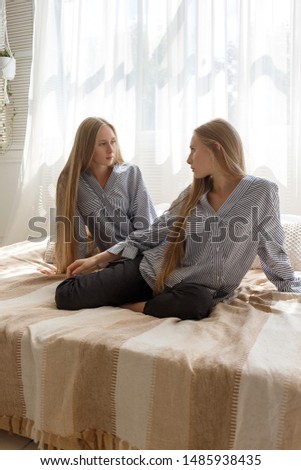 two pretty twin sisters in black jeans and striped oversize shirts with long blond hair sitting on sofa against background of window with curtain and looking at each other