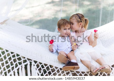 Cute little sister and brother having fun together in hammock in green summer garden, hugging and smiling. Happiness, childhood, summer time concept