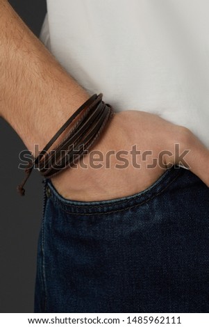 Cropped close-up shot of a male wrist with a multilayer bracelet with strings. The double-colored bracelet is made of brown wax ropes and black leather strips. The fashion model is dressed in jeans 