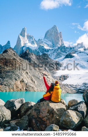 Couple in love & scenic view of snowcapped mountain tops of Mount Fitzroy, Patagonia trek. Blue sky, turquoise lake and scenic rock landscape. Shot in Argentina. Nature, travel, adventure, hiking.