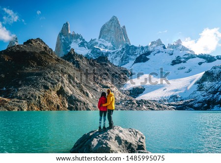 Couple in love & scenic view of snowcapped mountain tops of Mount Fitzroy, Patagonia trek. Blue sky, turquoise lake and scenic rock landscape. Shot in Argentina. Nature, travel, adventure, hiking.