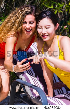 Two interracial young girls in bathing suits watching pictures together on the mobile phone, having fun and drinking beer sitting on sun loungers by a pool