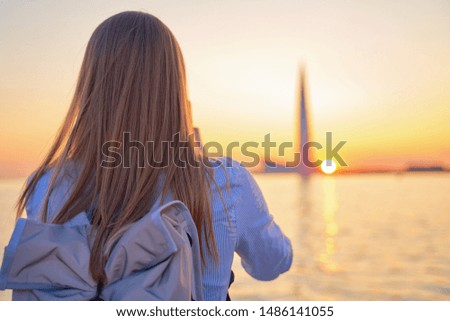 Young pleasant looking student with long dark loose hair, wearing casual T shirt and stylish grey backpack, making beautiful picture of sunset, sea and city sights. View from back.  