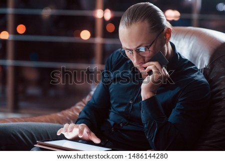 Credit card in the hand and tablet on the legs. Stylish businessman in eyewear works alone in the office at nightime.