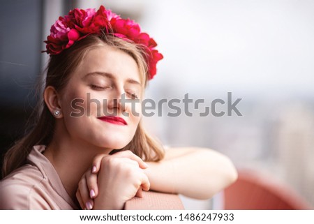 Close up portrait of young pregnant woman in beige dress and pink flower crown looking happy while sitting on the balcony. Horizontal shot. Selective focus