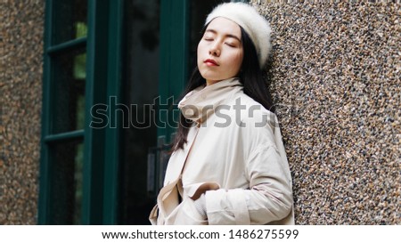 Beautiful young brunette woman in white beret and coat leaning against wall with eyes closed and hands in pocket. Street photography of a fashionable tired Chinese woman.