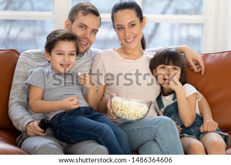 Happy family mom dad and cute little kids son daughter eat popcorn hold remote control watch tv show sit on sofa together, parents having fun lounge with children view television at home on couch