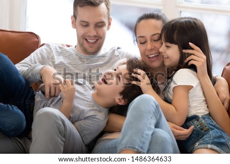 Cheerful funny family young parents couple and cute small children son daughter laughing bonding playing together on couch, happy mum dad having fun with kids tickling cuddling relaxing on sofa