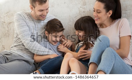 Funny happy family of four young parents couple and cute small children son daughter laughing playing sit on floor, carefree kids siblings bonding tickling cuddling with mom dad together at home