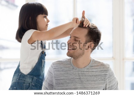 Cute funny little kid daughter playing with young single father brushing hair, small child girl holding comb pretend hairdresser doing hairstyle having fun with happy dad bonding together at home