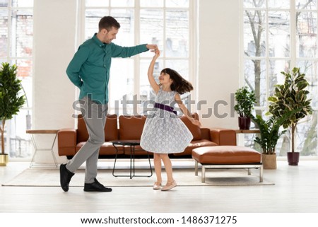 Loving young dad dancing with happy cute little daughter holding hand of kid girl princess wearing dress, caring father teaching waltz small child playing in living room enjoy time together at home