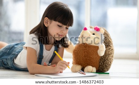 Cute happy little kid girl playing drawing pencils with fluffy hedgehog lying on warm heated floor at home, funny creative preschool small child teaching stuffed toy coloring picture having fun alone