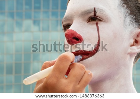 Street makeup. A close-up of the face of a teenager who draws an image of a clown or mime against the background of a modern city building. Preparing for the carnival for All Saints Day or Halloween.