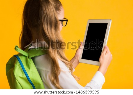 Educational Apps For School. Elementary Student Girl Looking At Blank Tablet Screen On Yellow Studio Background. Rear View, Mockup