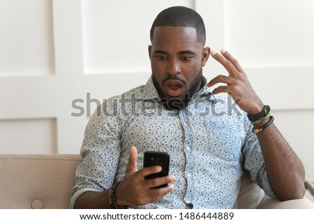 Surprised shocked millennial african american man holding phone, received mail with unbelievable news, amazing deal offer, grand sale discounts, new app free download, sitting on couch at living room. Royalty-Free Stock Photo #1486444889