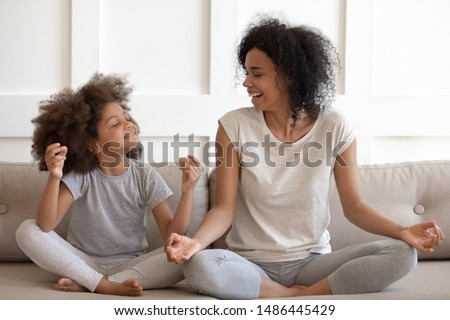 Excited african american woman teaching cute little adorable daughter meditating and practicing yoga exercises. Happy mixed race family sitting on comfy couch in lotus position, having fun together. Royalty-Free Stock Photo #1486445429