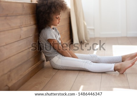 Upset african american cute kid girl sitting alone on heated warm floor in living room, feeling bored. Sad small adorable mixed race child feeling lonely, sleepy at home. Unhappy children concept. Royalty-Free Stock Photo #1486446347