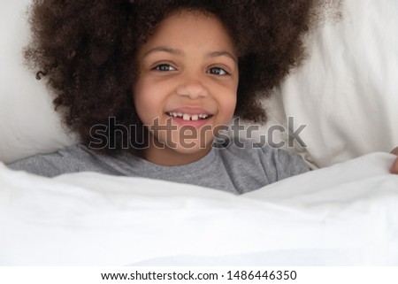 Top above view happy african american kid girl lying on pillow in bed under blanket, waking up in the morning after good dream, full of energy. Smiling mixed race adorable child looking at camera.