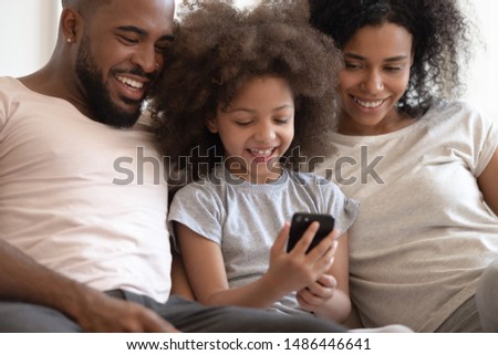 Close up headshot cropped image happy african american parents sitting together, watching little cute smiling kid daughter playing online game on smartphone or watching cartoons. Parental control.