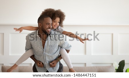 Excited african american man giving piggyback ride to happy cute small daughter, enjoying active leisure time. Laughing black father carrying on back little baby girl playing, having fun together. Royalty-Free Stock Photo #1486447151