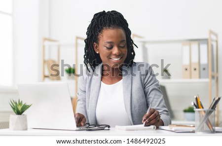 Smiling Afro Businesswoman Working On Laptop And Taking Notes In Modern Office. Free Space Royalty-Free Stock Photo #1486492025