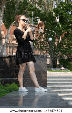 Girl learning to play trombone. Girl plays standing on the alley of a city park. Portrait.