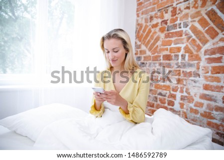 Beautiful blonde woman wearing pajama using phone in her bedroom.