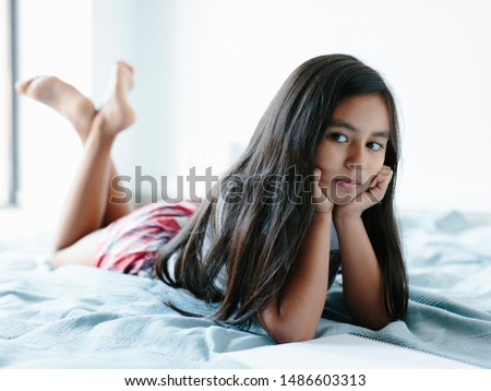 A 9-year old girl is lying on a blanket on bed. She's looking to her right. She has long dark hair. She has hispanic ethnicity features. She rests her head on her hands, looking bored or upset.