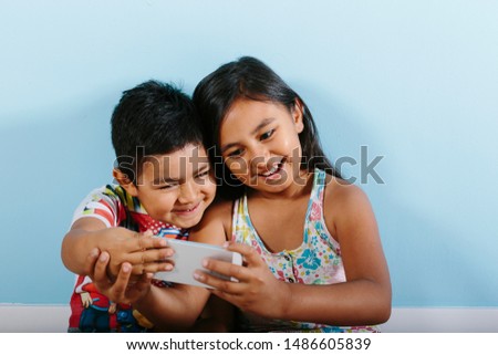 Two siblings, a 9-year old girl and a 5-year old boy are taking a selfie with a smartphone. They're smiling.They're leaning over a blue wall. They are wearing summer casual clothes.