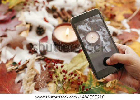 Hand holding phone and taking photo of autumn flat lay. Candle, berries, fall leaves, anise,herbs, acorns, nuts, cinnamon, cotton on white textile. Hygge lifestyle, cozy autumn mood