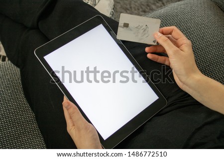 Young woman with a credit/bank card about to pay for something on her tablet sat on a sofa, the tablet has a white full size screen to enable adding text or images. 