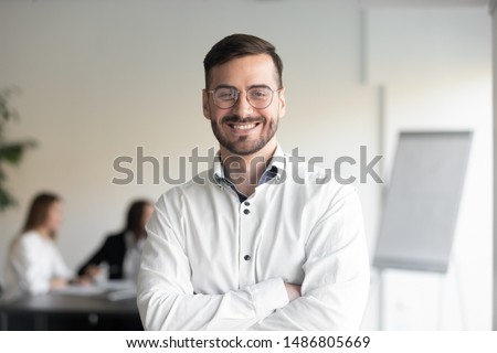 Confident smiling business man professional coach wear glasses stand arms crossed looking at camera, happy male executive company owner corporate manager leader in office, headshot close up portrait