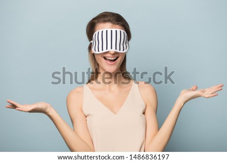 Happy woman wearing sleeping mask on head isolated on blue studio background feel positive excited, smiling young female model in homewear demonstrate advertise beauty product show it on face