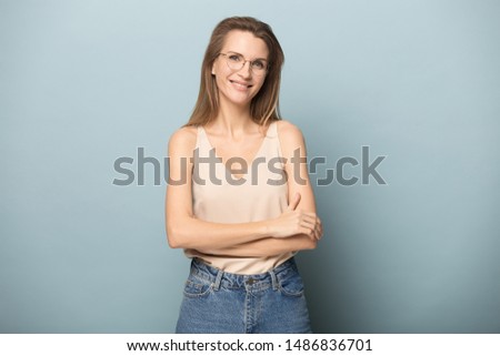 Portrait of smiling beautiful blonde woman wear glasses stand isolated on blue studio background with hands crossed, happy pretty female model in casual clothes and spectacles pose looking at camera