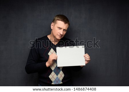 Studio waist-up portrait of focused caucasian fair-haired man wearing sweater, holding white blank paper sheet with place for your text, looking seriously at camera, over gray background