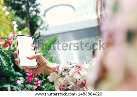 woman's hand holding and using smartphone or tablet with blank white screen in modern garden of shopping mall, shallow depth of field
