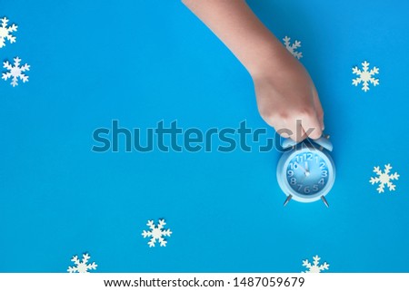 Child's hand holding blue alarm clock showing five to twelve with paper snowflakes. New Year's night, midnight. Flat lay concept image, top view on pastel blue paper background with copy-space.