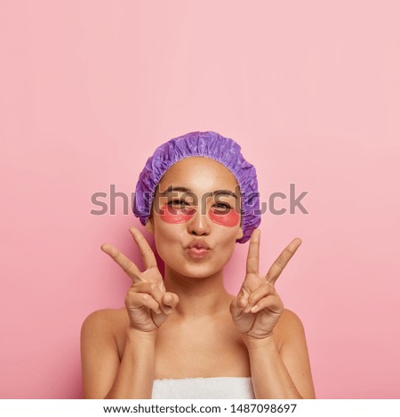 Beauty and rejuvenation concept. Pretty korean woman makes peace hand gesture, keeps lips folded, has under eye patches on face, wears purple bath hat, enjoys spa procedures after taking shower