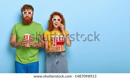 Happy ginger woman and sad man wears cinema glasses, watch film premier, eat delicious popcorn together, stand closely to each other against blue background, free space aside. Spare time concept