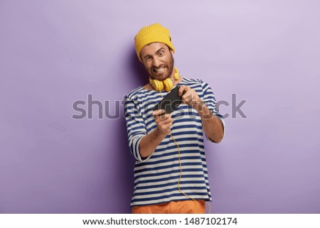 Handsome unshaven man holds smartphone horizontal, enjoys new awesome application, plays game online, has fun indoor wears yellow hat and sailor jumper struggles to win competition gains highest score