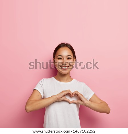 Attractive Asian female makes heart shape gesture, expresses love, says be my valentine, smiles positively, wears white outfit, poses against pink wall with empty space. Body language concept