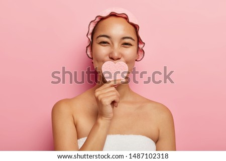 Beautiful satisfied korean female holds sponge in shape of heart, washes face, cleans skin, takes care of her complexion, has beauty spa treatment, stands in bathroom wrapped in towel, wears showercap Royalty-Free Stock Photo #1487102318