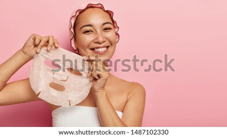 Attractive young chinese woman applies paper sheet mask on face, enjoys beauty skin care, stands wrapped in towel, wears bath cap, smiles positively, isolated on pink background. Rejuvenation concept Royalty-Free Stock Photo #1487102330