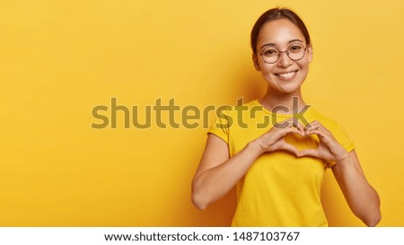 Lovely korean woman with happy smile, shapes heart with both hands, expresses love to you, wears round spectacles and yellow t shirt, says be my valentine flirts with boyfriend poses indoor copy space