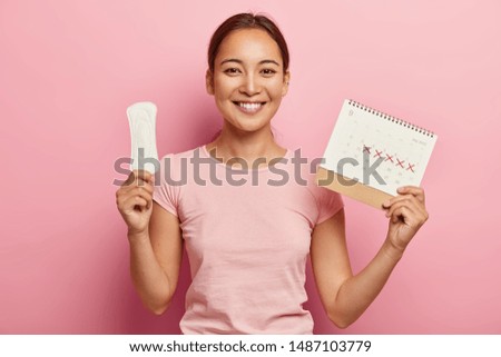 Adorable dark haired korean woman holds clean pad and menstruation calendar, has pleased face expression, happy to have regular menstrual cycle, wears pink t shirt. Women, hygiene, health care concept