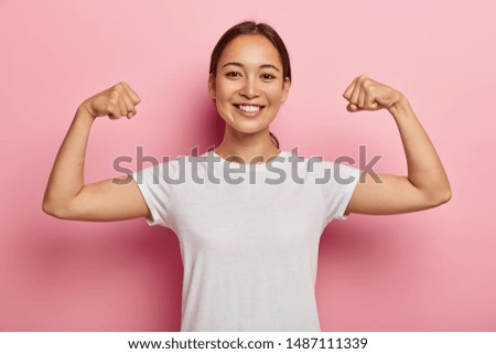 Pretty korean female model keeps fit and healthy, raises hands and shows muscles, feels proud about her achievements in gym, smiles broadly, dressed in white casual wear, poses indoor shows real power Royalty-Free Stock Photo #1487111339