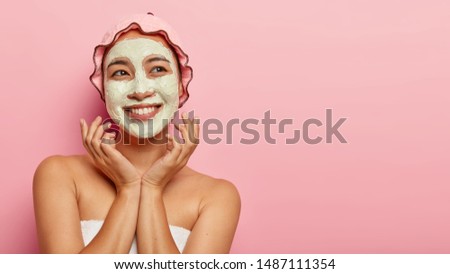 Face care concept. Positive lovely woman has clean fresh skin, applies rejuvenation clay mask on face, smiles positively, keeps hands near cheeks, wears protective shower cap, isolated on pink wall Royalty-Free Stock Photo #1487111354