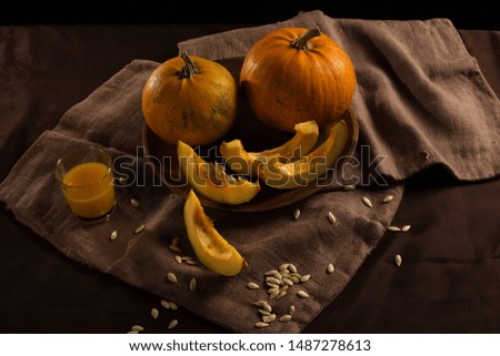 Two pumpkins and a glass of juice surrounded by slices and pumpkin seeds against a dark background